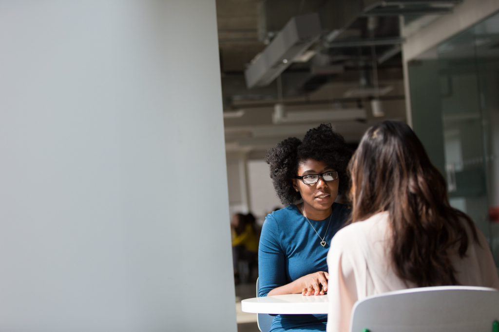 Dans une cafétéria, une femme se présente de dos et parle à une femme en face d'elle qui nous apparaît de face sur sa gauche. Elles sont toutes deux assises à une table.