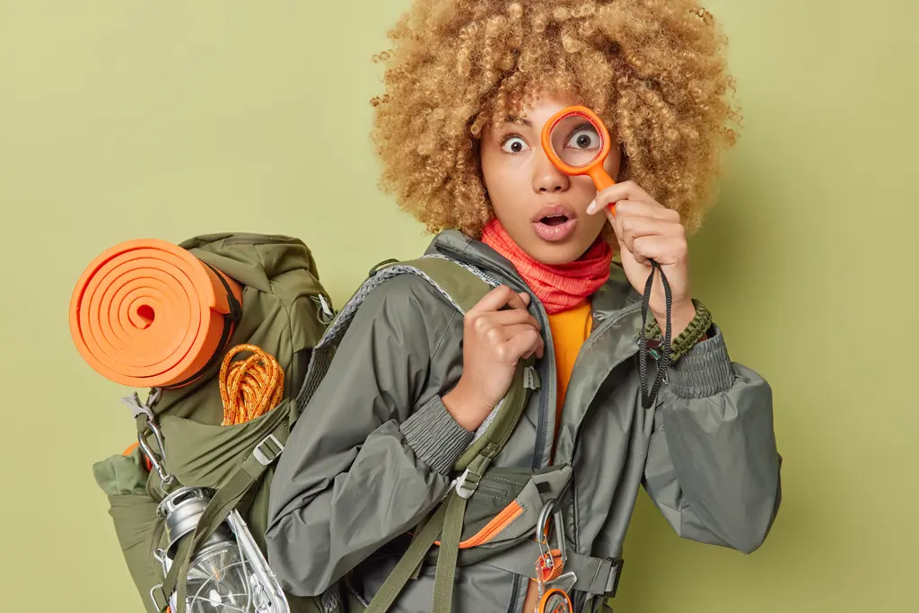Une femme en habit de randonné tient une loupe à son oeil gauche et regarde directement la caméra.